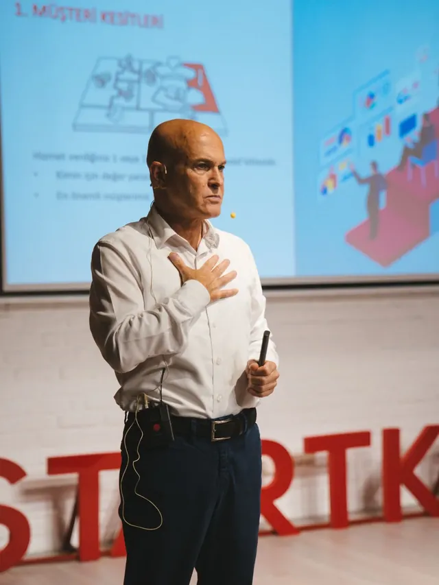 Güven Açık bir eğitim etkinliğinde sunum perdesinin önünde konuşuyor