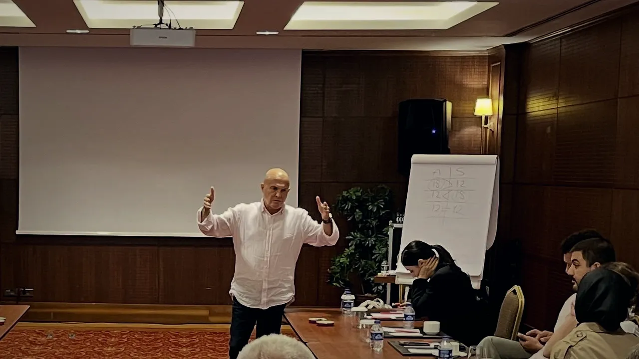 Güven Açık, kâğıt tahtadaki rakamların yanında anlatıyor