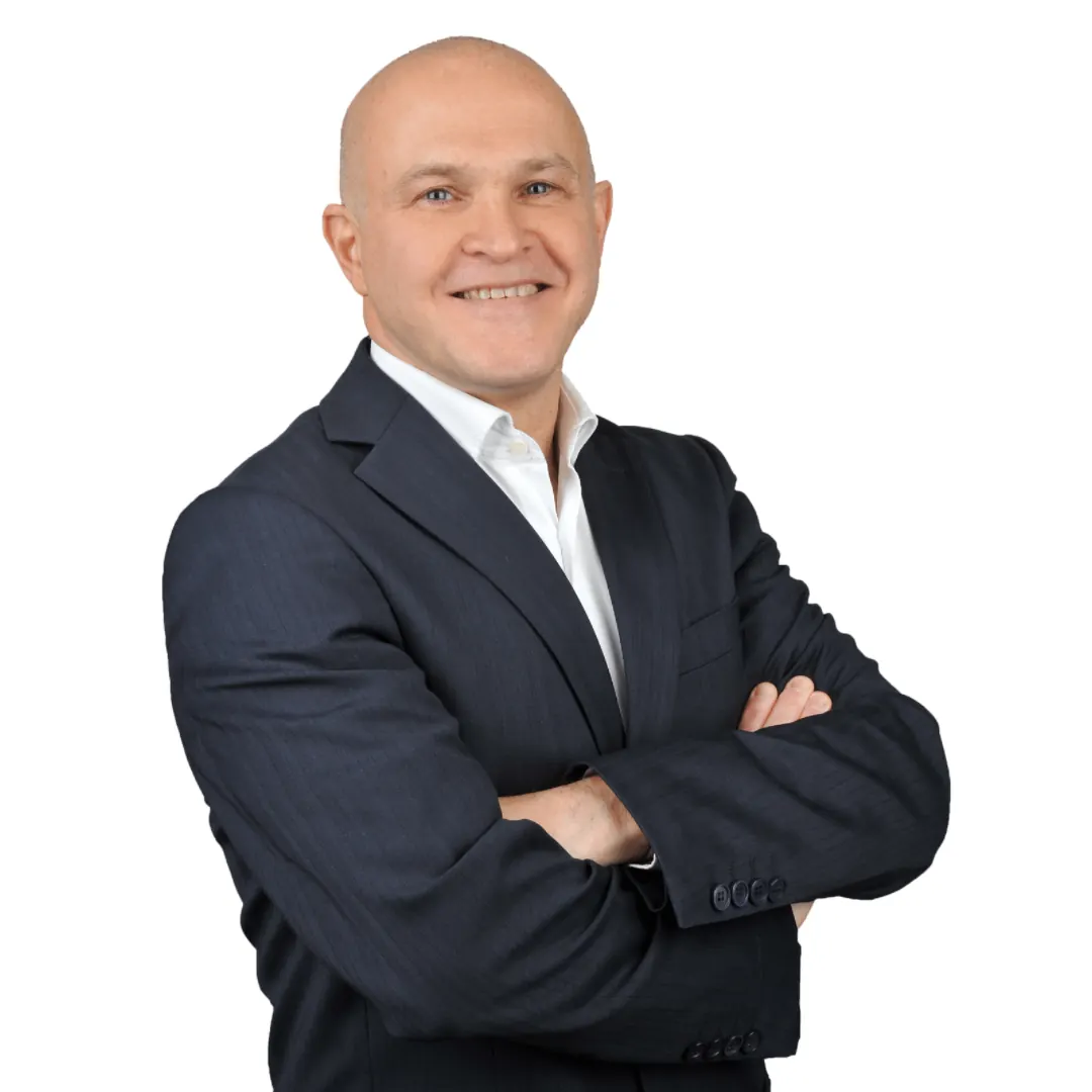 Koyu renk takım elbiseli, kollarını kavuşturmuş bir adam beyaz fonun önünde gülümsüyor
