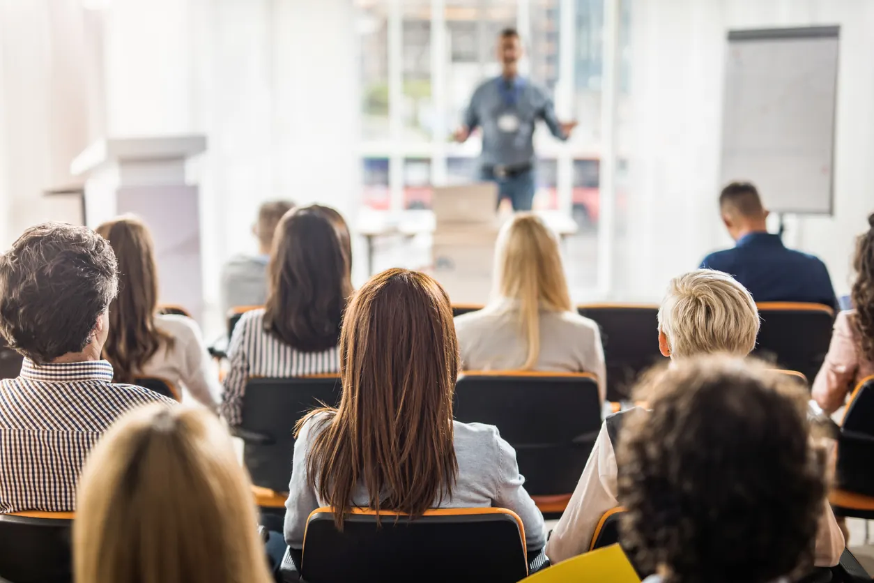 Seminer salonunda oturan katılımcılar karşılarında ayakta konuşan eğitmeni dinliyor