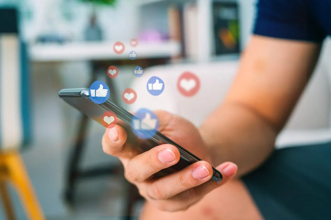 Akıllı telefon tutan bir elin üzerinde beğeni ve kalp simgeleri uçuşuyor