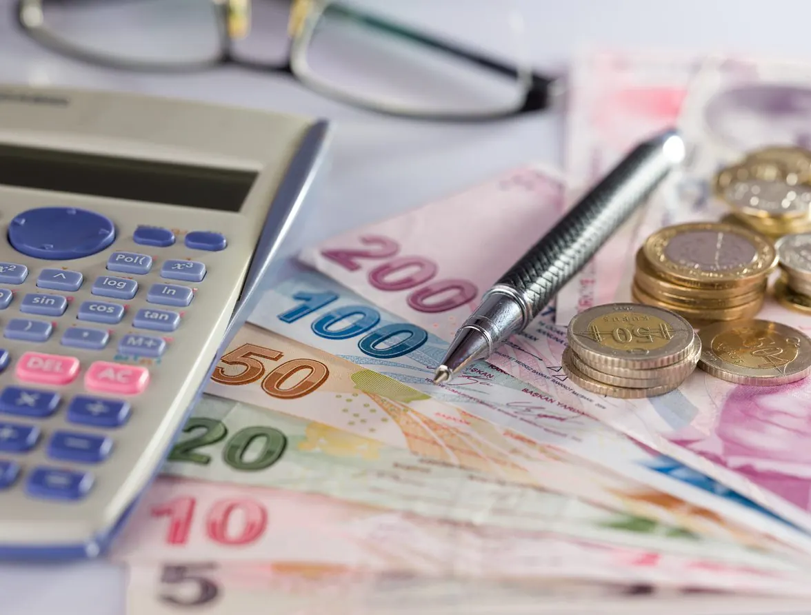 Masada hesap makinesi, kalem, gözlük, Türk lirası banknotları ve madeni paralar duruyor