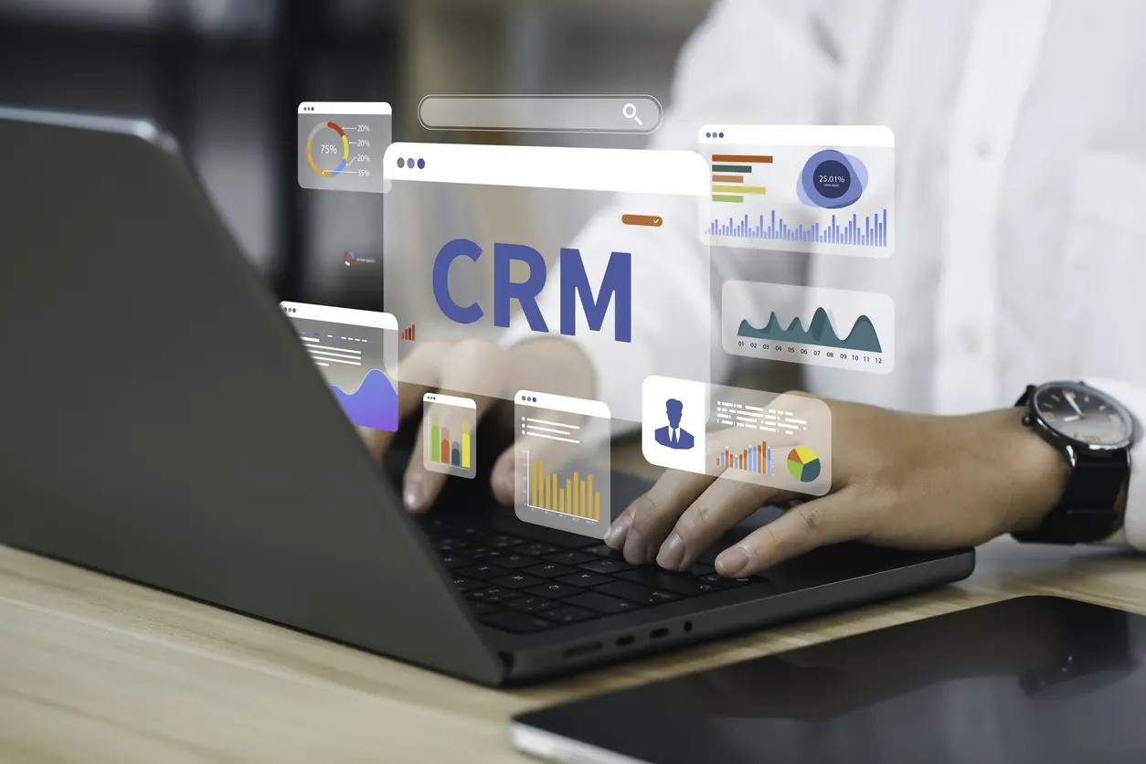 Dizüstü bilgisayarda yazı yazan ellerin üzerinde “CRM” yazısı ve grafik panoları görünüyor