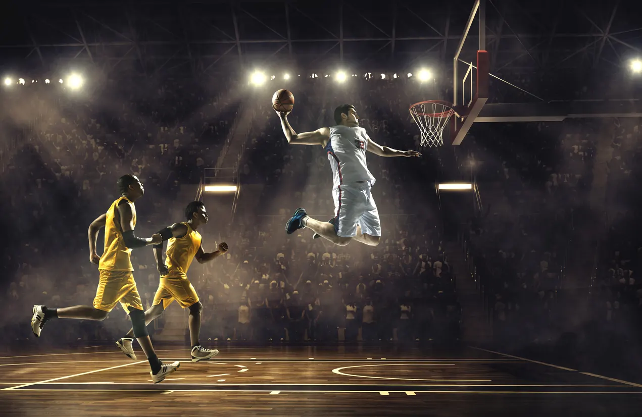 Basketbol sahasında beyaz formalı oyuncu potaya sıçrıyor, sarı formalı iki rakibi arkasından koşuyor
