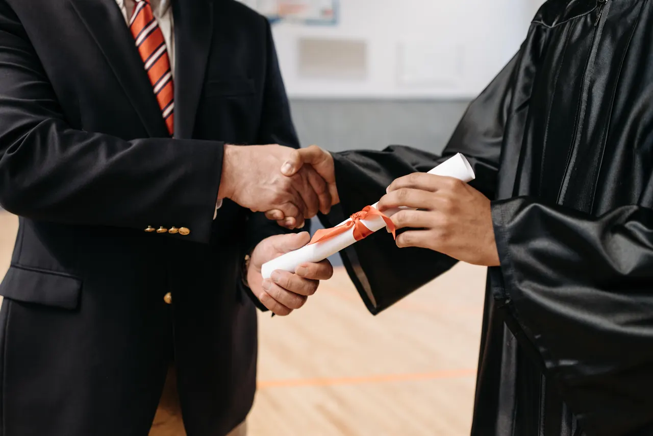 Takım elbiseli adam, cübbeli mezunun elini sıkarak kurdeleli diplomayı veriyor
