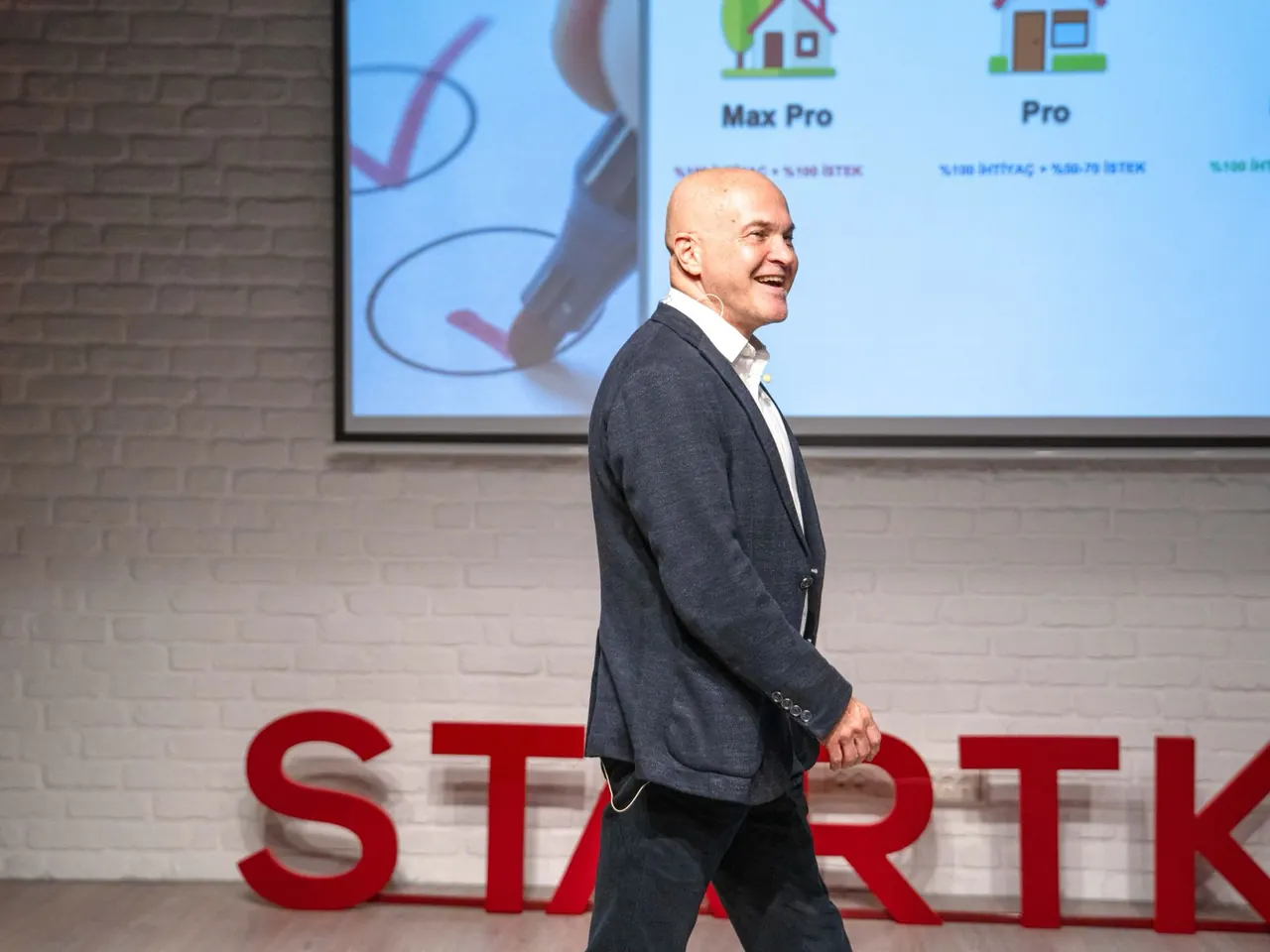 Güven Açık sahnede gülümseyerek yürüyor, arkasında sunum perdesi ve kırmızı Startkey harfleri var